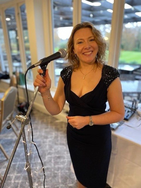 Sarah Locke stood at a microphone in a care home in a black evening dress during a wellbeing performance